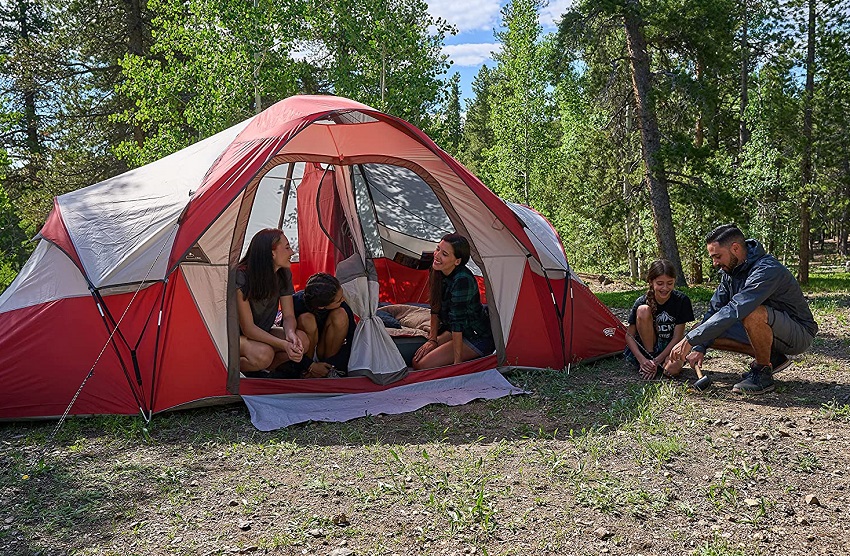 A man and a girl finish pitching the Wenzel Bristlecone 8-Person Dome Tent, while three women already sit inside that tent and smile at each other
