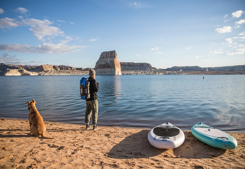 A woman with a dry bag, her dog, and two stand up paddleboards are on a lake shore