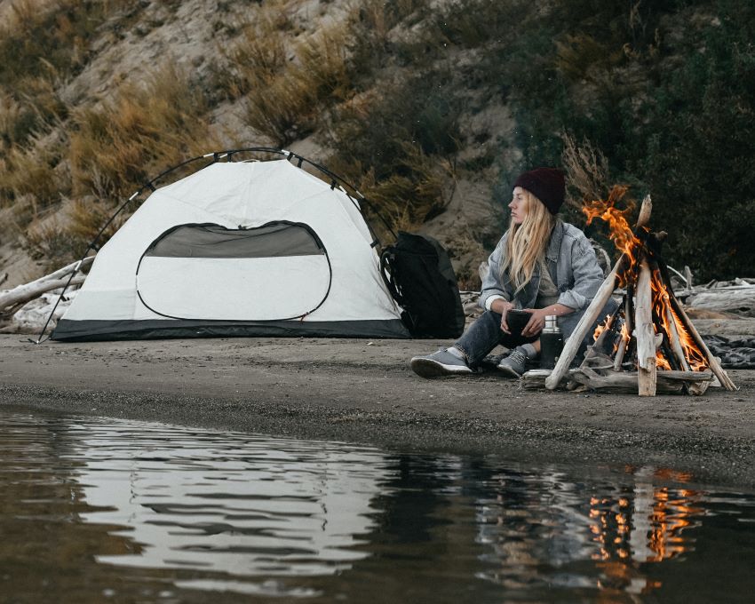 A woman sits near a campfire and a white tent on a lake shore