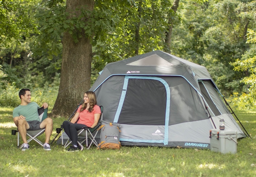 A man and a woman sit on camp chairs near the Ozark Trail 6-Person Dark Rest Instant Cabin Tent in the woods