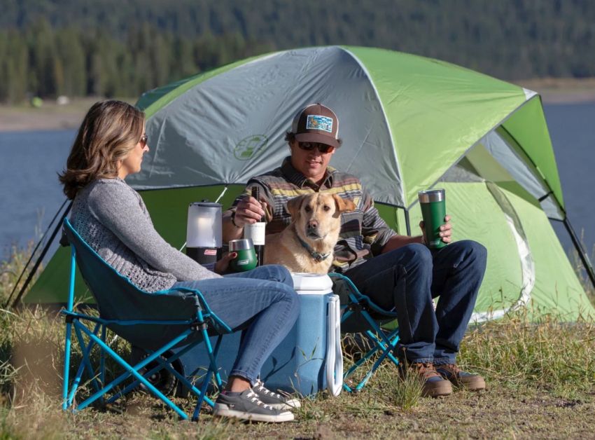 A man, a woman and a dog sit in camping chairs near the green Coleman Sundome 3-Person Camping Tent