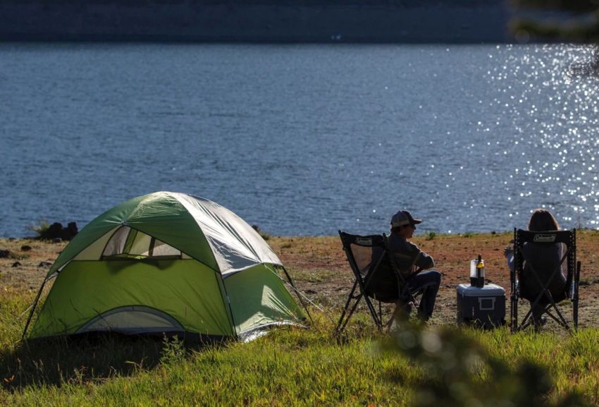 A man and a woman sit in camping chairs near the green Coleman Sundome 3-Person Camping Tent on a lake shore