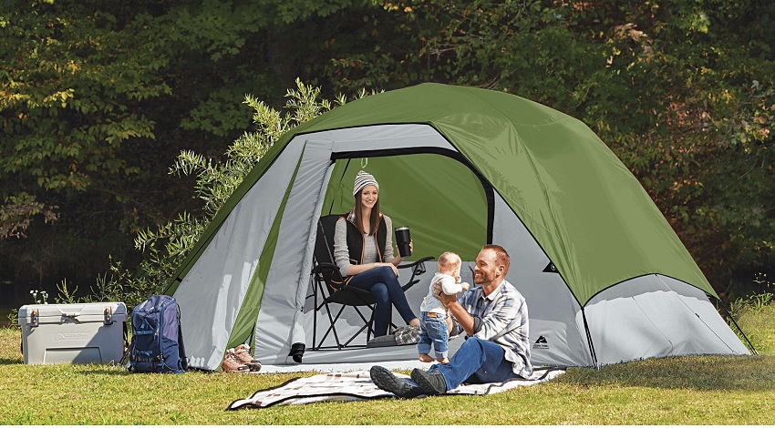 A woman sits inside the Ozark Trail 6-Person Clip & Camp Dome tent and looks at a man playing with a child outside.