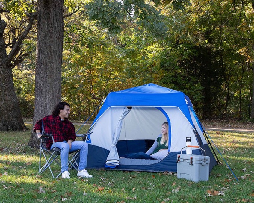 A man sits in a chair near the Ozark Trail 4-Person Instant Cabin tent with LED Lighted Hub and looks at a woman, sitting inside that tent
