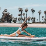 A woman practices yoga, sitting on a twine on her SUP