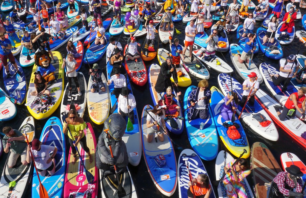 A crowd of people paddle SUPs of different shapes and colors