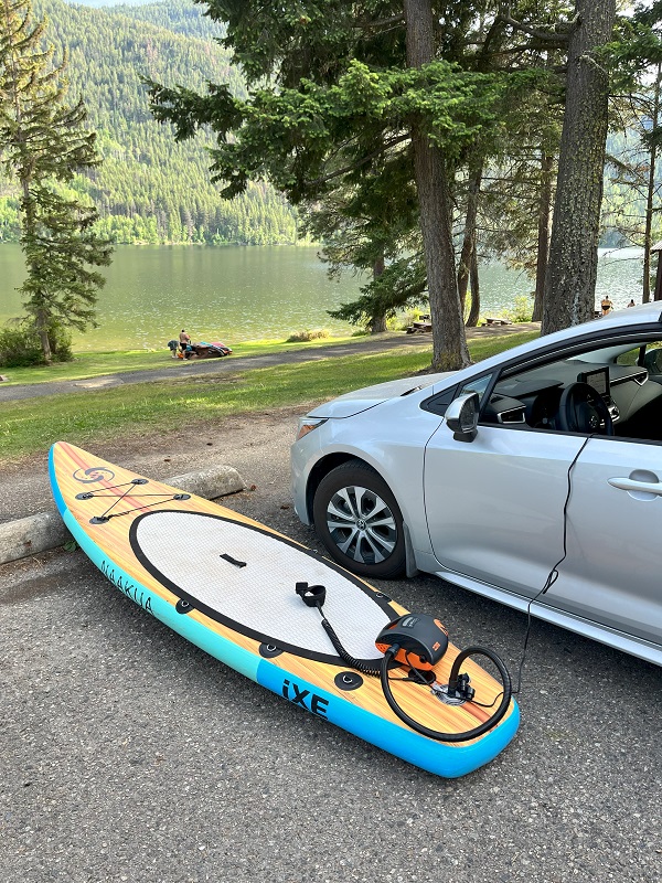 OutdoorMaster Shark II pump is connected to an inflatable SUP and the 12V port inside a gray car on a lake shore