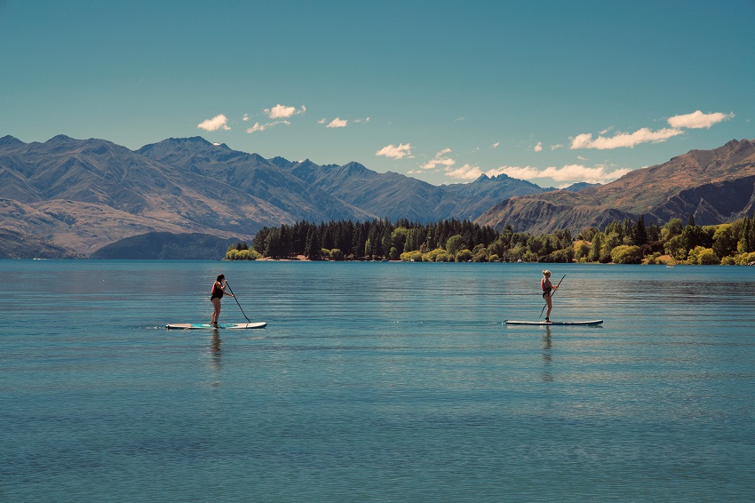 Two women paddle their SUPs on a lake, surrounded by mountains