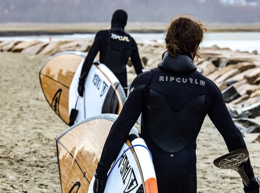 Two men in black wet suits carry their paddle boards towards water
